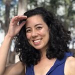 April has black curly hair and a big smile with dimples. She is wearing a dark blue collared shirt that compliments her pale olive skin and has a warm and friendly demeanor.