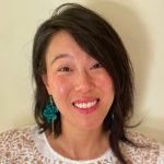 Juliette is a Korean American woman with long black hair. She is standing in front of a white wall and smiling, while wearing a white shirt with gold embroidery and green earrings shaped like a snake.