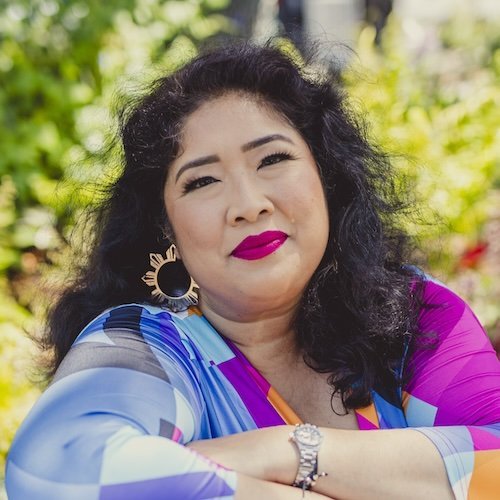 Sheilani is a filipina woman with medium-length, dark, curly hair. She is smiling and wearing a colorful dress while outdoors on a sunny day with trees in the background.
