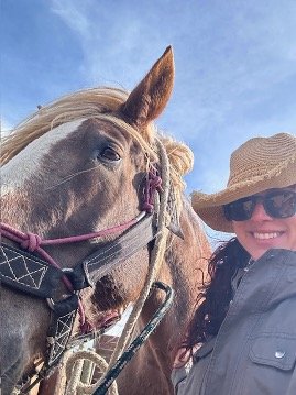 Kristi Chester Vance smiling in sunglasses and large brim hat with a draft horse