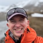 Bruce is a man wearing a baseball cap with sunglasses on it and an orange windbreaker, smiling with water and snowy mountains in a slightly out of focus background.