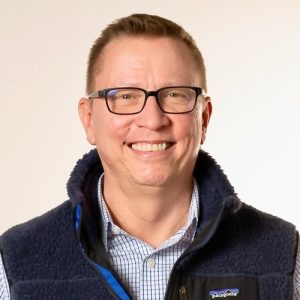 Matt is a white man with short brown hair and no facial hair. He's wearing a collared white and blue checked shirt under a dark blue fleece vest in this formal portrait with a beige background