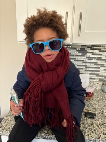 Caira's son Orion with blue sunglasses and a maroon scarf on sitting on the kitchen counter