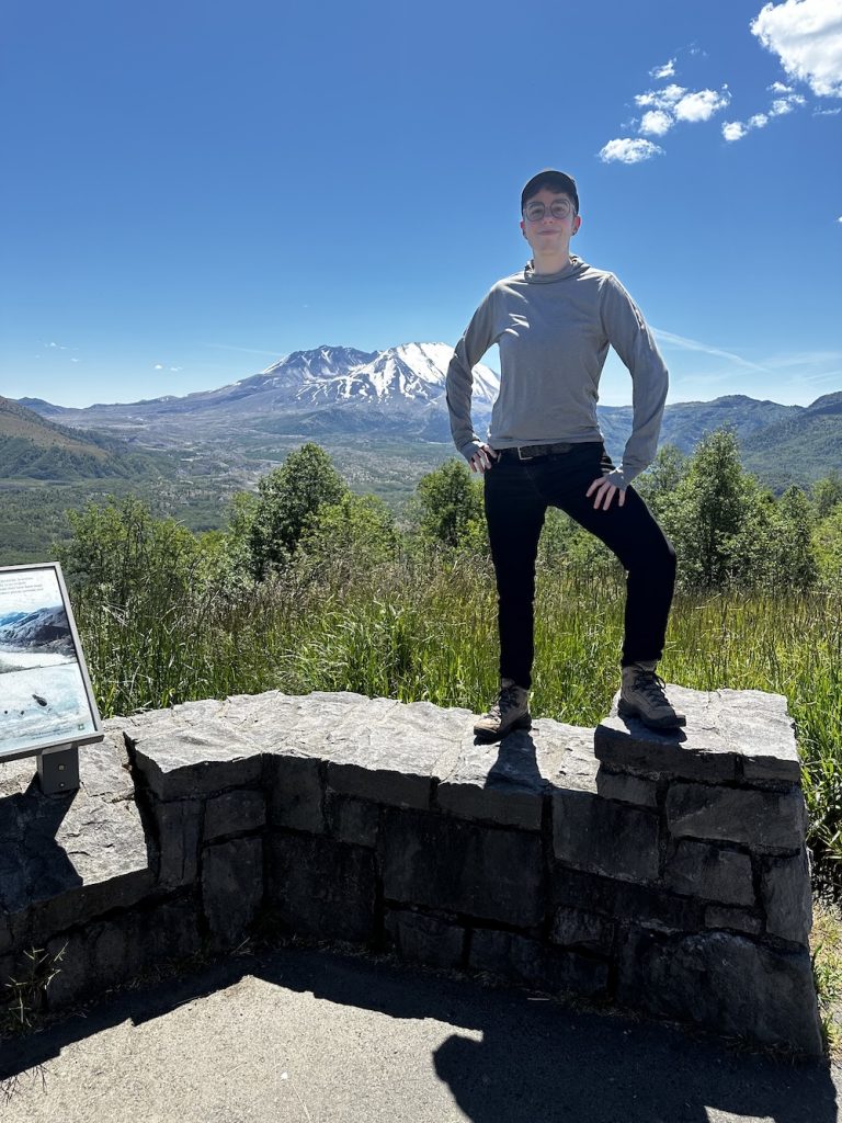 maia is standing on a rock wall in hiking gear with a big snowy mountain in the background