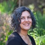 aparna is a South Asian woman with curly salt and pepper chin-length hair. A green shirt peaks out from the edges of her black cardigan sweater in this formal garden portrait, with sun-dappled plants in the background