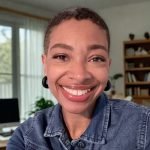 Kellie is a black woman with short hair, posing in this portrait with a big smile and wearing a jean jacket in her home office.