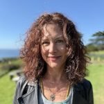 Kristi is a white woman with really curly reddish hair. She’s wearing a black leather jacket and a silver necklace. She’s smiling and standing on a green lawn with blue sky in the background.