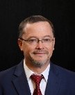 Man with short borwn hair and beard wearing clear framed glasses and a suit with a red tie in this formal protrait