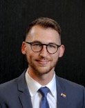 Man with brown hair and a short beard, wearing glasses and a suit in this formal portrait